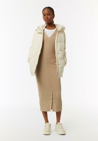 Cremfarbener Pufferjacke mit Kapuze, getragen über einem beige gerippten Strickkleid mit Knöpfen, kombiniert mit cremfarbenen Turnschuhen.