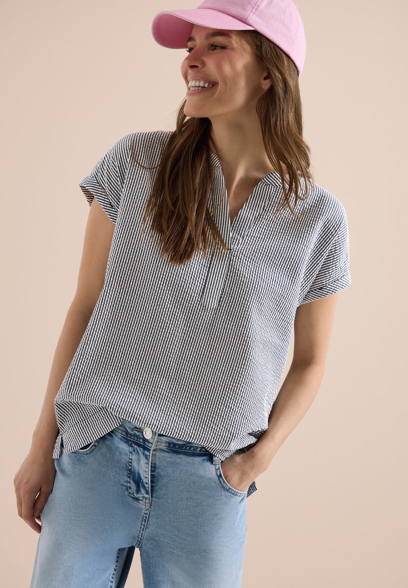 Femme portant une casquette rose, un t-shirt rayé à manches courtes et un jean bleu clair, souriante et regardant sur le côté avec une main dans sa poche.