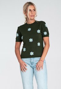 Blondynka w ciemnozielonym krótkim swetrze z jasnoniebieskimi wzorami w kwiaty oraz jasnoniebieskich dżinsach, patrząca w bok.