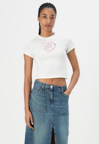 Balta īsā T-kreklā ar rozā "TOMMY JEANS" grafiku, kombinēta ar zilu džinsu svārku ar priekšējo šķēlumu un tradicionālajiem kabatām.