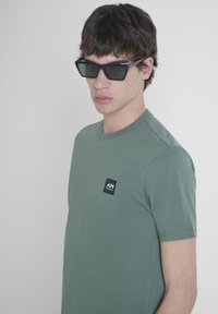 T-shirt verde a maniche corte con collo rotondo e una piccola etichetta nera con testo bianco sul petto sinistro. Il tessuto sembra liscio.