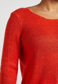 Roter, gestrickter Pullover mit loser Maschenstruktur, weitem Rundhals-Ausschnitt und langen Ärmeln. Weiche Textur mit einem leichten Glanz.