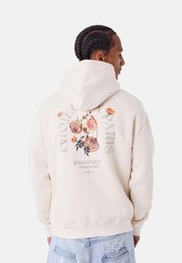 Sudadera con capucha crema que presenta bordados florales en la parte posterior, con el texto "PROJECT PARIS WILD SPIRIT PXP COLLECTION."