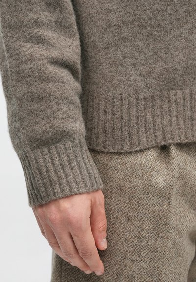 Maglione marrone in lana con polsini e orlo a coste, che mostra una morbida texture. Mano visibile, con pantaloni beige e testurizzati sullo sfondo.