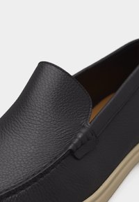 Mocassin en cuir noir avec surface texturée et semelle en caoutchouc beige, montrant des détails en gros plan de dessus et de côté.