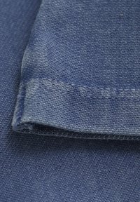 Denim w głębokim niebieskim odcieniu, ukazujący teksturowany splot z widocznym przeszyciem i zagiętą krawędzią. Gładka powierzchnia z niewielkimi odchyleniami.