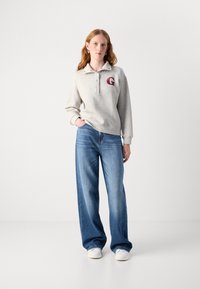Grå sweatshirt med krage och knappslå, med en röd "G"-logotyp. Matchad med vida blå jeans och vita sneakers.