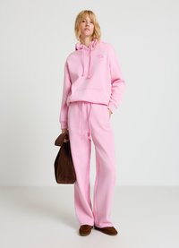 Sweat à capuche en coton rose avec cordons de serrage et poche avant, assorti à un pantalon de survêtement rose. Sac en suède marron et chaussures marron complètent le look.