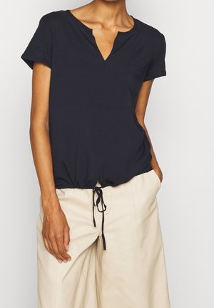 Navyblaues Kurzarm-Top mit V-Ausschnitt, gerafftem elastischen Saum und seitlichen Taschen. Hergestellt aus weichem Stoff mit glatter Textur. Dazu werden helle Hosen getragen.