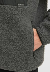 Graue Fleecejacke mit einer strukturierten, weichen Oberfläche, ausgestattet mit einer Reißverschlusstasche, schwarzen Akzenten und einem Etikett, das "11 PROJECT" liest.