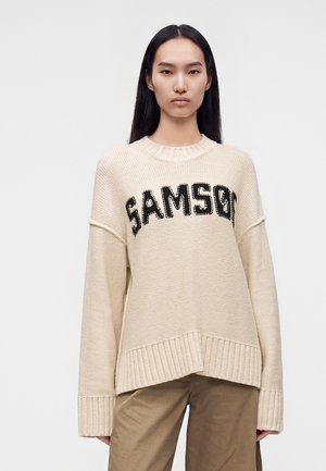 Jeune femme aux longs cheveux noirs portant un pull beige en maille avec le texte "SAMSO" et un pantalon marron sur un fond clair et uni.