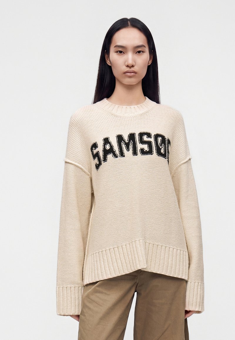 Jeune femme aux longs cheveux noirs portant un pull beige en maille avec le texte "SAMSO" et un pantalon marron sur un fond clair et uni.