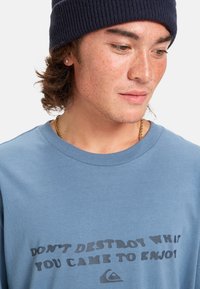 Camiseta de algodón azul claro con un diseño de texto negro ondulado que dice: "NO DESTRUYAS LO QUE VINISTE A DISFRUTAR", acompañado de un logo.