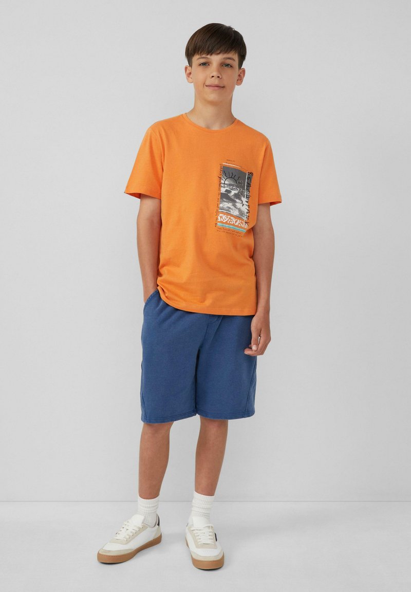 Oranje katoenen T-shirt met een grafisch zakontwerp, gecombineerd met marineblauwe shorts. Witte sneakers met beige accenten en witte sokken maken de look compleet.