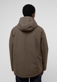 Giacca marrone impermeabile con cappuccio, caratterizzata da una texture liscia, una forma leggermente svasata e un logo sul manica sinistra.