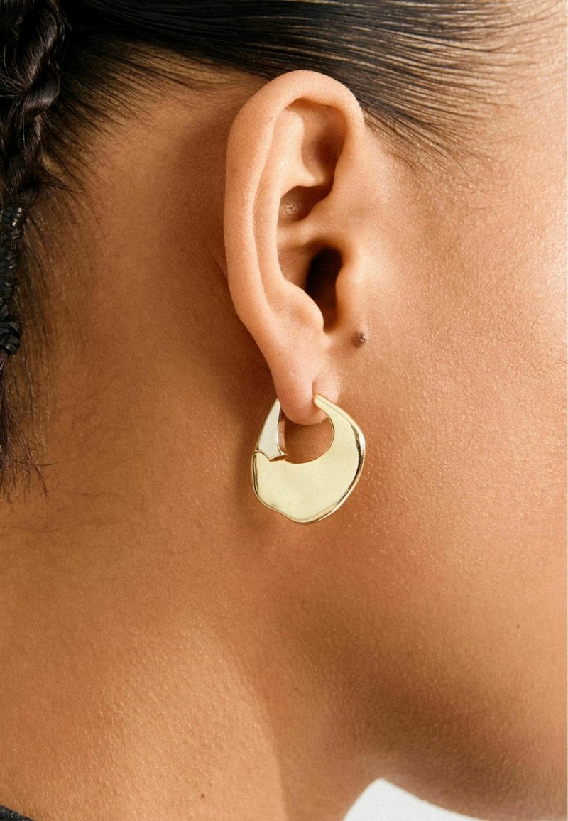 Primo piano di un orecchio di una persona che indossa un brillante orecchino a cerchio in oro di forma irregolare, con capelli scuri intrecciati visibili dietro l'orecchio.