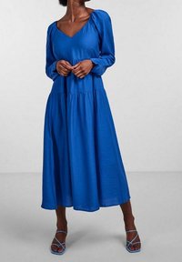 Vestido azul de manga larga con escote en V, tejido texturizado, falda acampanada y mangas abullonadas elásticas. Combinado con sandalias azul con tiras.