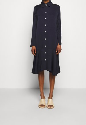 Person trägt ein langärmeliges, marineblaues Hemdkleid mit ausgestelltem Saum und beigen offenen Schlupfschuhen vor einem einfarbigen Hintergrund.