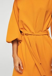 Pomarańczowa sukienka z długim rękawem, z dopasowaną talią i paskiem do przewiązania. Miękka, strukturalna tkanina o luźnym kroju oraz marszczonych rękawach.