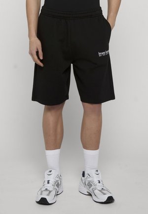 Person trägt schwarze knielange Shorts mit dem Text "bruno banani", weiße Sportsocken und weiße New Balance Turnschuhe, steht mit den Händen in den Taschen.