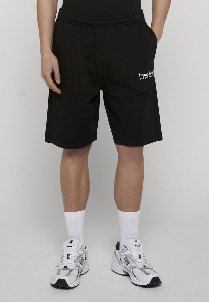 Personne portant un short noir jusqu'aux genoux avec le texte "bruno banani", des chaussettes blanches montant jusqu'à la cheville et des baskets blanches New Balance, debout avec les mains dans les poches.