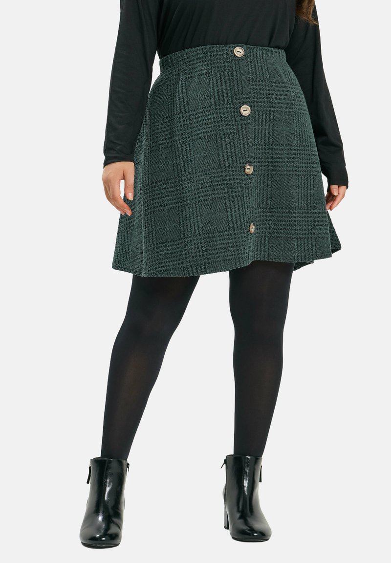 Grüne Karotte aus strukturierter Stoff, mit einer ausgestellten Form und einem vierknöpfigen Frontverschluss. Getragen mit schwarzen Strumpfhosen und Stiefeletten.