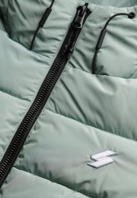 Lys grøn polstret jakke med en sort lynlås, elastiske snore og et lille hvidt broderet logo på stoffet. Struktur overflade.