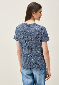 Maglietta blu navy con un motivo astratto ondulato bianco, maniche corte e scollatura a girocollo, abbinata a jeans in denim blu chiaro.