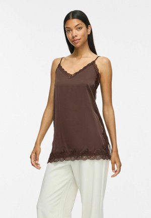 Femme aux longs cheveux foncés portant un haut sans manches marron orné de dentelle et un pantalon blanc ample, debout avec une expression neutre.