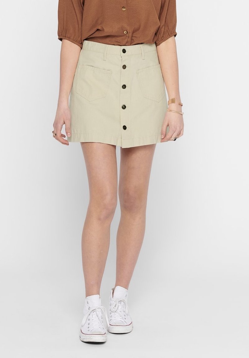 Gonna di denim beige con chiusura frontale a bottoni, due tasche anteriori e lunghezza a metà coscia. Abbinata a sneakers bianche e un top marrone.