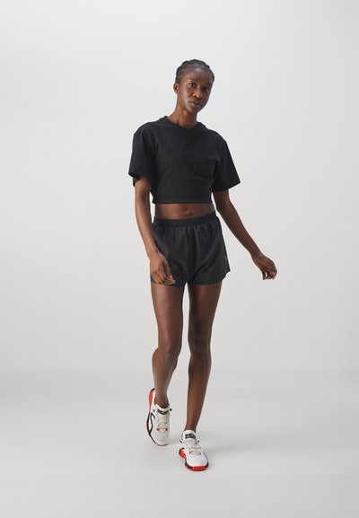 Frau in schwarzem, kurzem Crop-T-Shirt und Shorts, die nach vorne geht und weiße und orangefarbene Laufschuhe auf hellgrauem Hintergrund trägt.