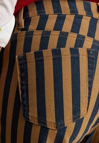 Blugi din denim cu dungi verticale alternative în nuanțe de maro deschis și albastru închis. Include un buzunar posterior cu detalii vizibile de cusătură.