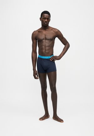 Atletski muškarac stoji bos s jednom rukom na kuku, nosi mornarsko plave bokserice sa svijetloplavim pojasom.