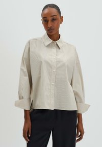 Beige button-up shirt with subtle vertical stripes, oversized fit, and cuffed sleeves. Made from smooth fabric with a classic collar design.