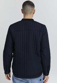 Giacca trapuntata blu navy con colletto a coste, caratterizzata da linee testurizzate e polsini con bottoni. Design minimalista con una finitura liscia.