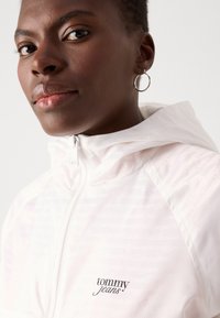 White hooded jacket with a shiny, lightweight material featuring an all-over tonal logo pattern. Small black "tommy jeans" logo on chest.