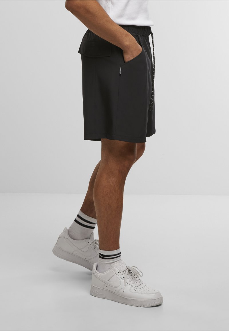 Person, der schwarze knielange Shorts mit Seitentaschen, weiße Socken mit schwarzen Streifen und weiße Nike-Sneaker trägt und seitlich steht.