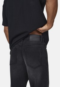 Czarne dżinsy z denimu o gładkiej fakturze, z dwoma tylnymi kieszeniami i skórzaną naszywką na pasku. Prosta czarna koszulka częściowo je zakrywa.