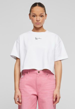 Tricou scurt alb cu mâneci scurte, având un logo brodat negru pe piept. Purtați cu o pereche de pantaloni din denim cu talie înaltă, roz.