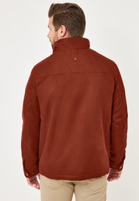 Rostfarbende Jacke mit gestepptem Schulterbereich, geknöpften Manschetten und hohem Kragen. Hergestellt aus strukturiertem Material, verfügt sie über einen Rückenkeil für eine optimale Passform.