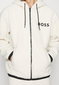 Cremefarbener Fleece-Kapuzenpullover mit Reißverschluss, schwarzen Akzenten und Kordelzügen. Verfügt über Seitentaschen und das schwarze "BOSS"-Logo auf der Brust.