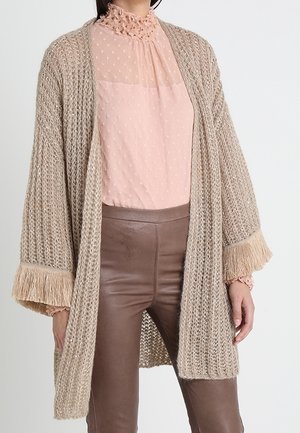 Femme portant un cardigan en grosse maille beige avec des poignets à franges par-dessus un chemisier rose transparent et un pantalon en cuir marron.