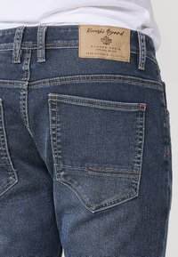 Jeans de mezclilla en azul oscuro con una textura rugosa. Las características incluyen costuras amarillas, una etiqueta de marca de piel y costuras en rojo en los bolsillos.