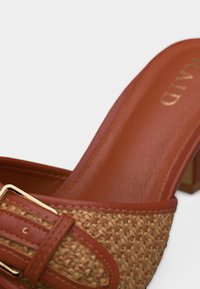 Mule en cuir marron avec détail en paille tissée et boucle dorée sur le talon bloc, intérieur de la chaussure visible.