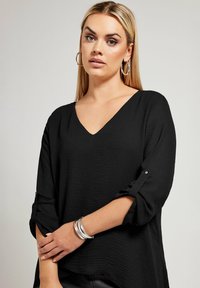 Blusa nera a V con maniche a tre quarti arrotolate e tessuto texturizzato. Apparentemente leggera, presenta una vestibilità morbida e polsini con bottoni.