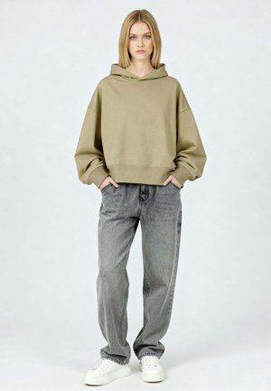 Jonge vrouw staat naar voren gericht, draagt een oversized olijfgroene hoodie, losse grijze jeans, witte sneakers, handen in de zakken, neutrale achtergrond.