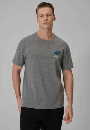 Mężczyzna w szarej koszulce z krótkim rękawem z logo „4F All-Round Sports Apparel” na piersi, stojący na jednolitym szarym tle.