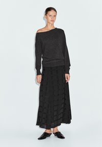 Maglione grigio scuro off-shoulder con maniche lunghe, abbinato a una gonna maxi plissettata nera con motivo a griglia, indossata con scarpe slip-on nere.
