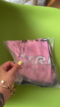 Rožinė sportinė marškinė, sulankstyta ir uždaryta aiškiame plastiko maišelyje, su ryškiu juodu logotipu ir tekstu. Rankoje laikomas paketas.