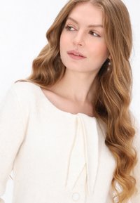 Pulover de culoare crem, cu un decolteu rotund, având o textură moale și un accent cu fundiță la guler, prins cu un nasture.